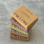 Four Dillon soap bars of different colors stacked on a textured gray surface