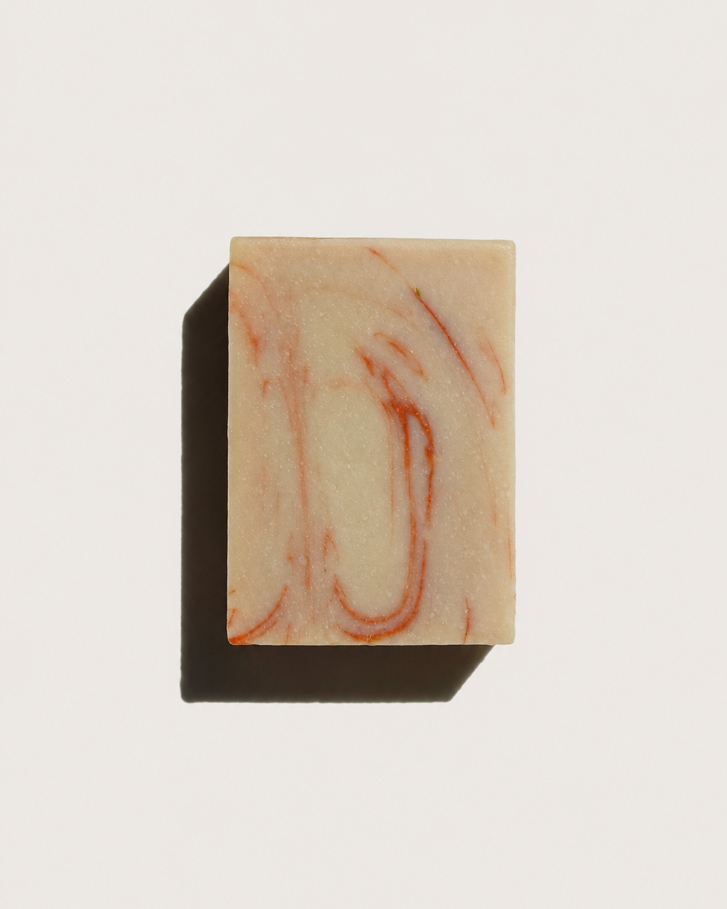A rectangular bar of soap with a cream-colored base and reddish-orange marbled swirls, casting a dark shadow on a plain white background.