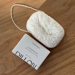 A white textured konjac body cleansing sponge next to bar soap in white paper packaging, labeled 'DILLON PURE MILK CLEANSING BAR', placed on a light brown wooden surface.