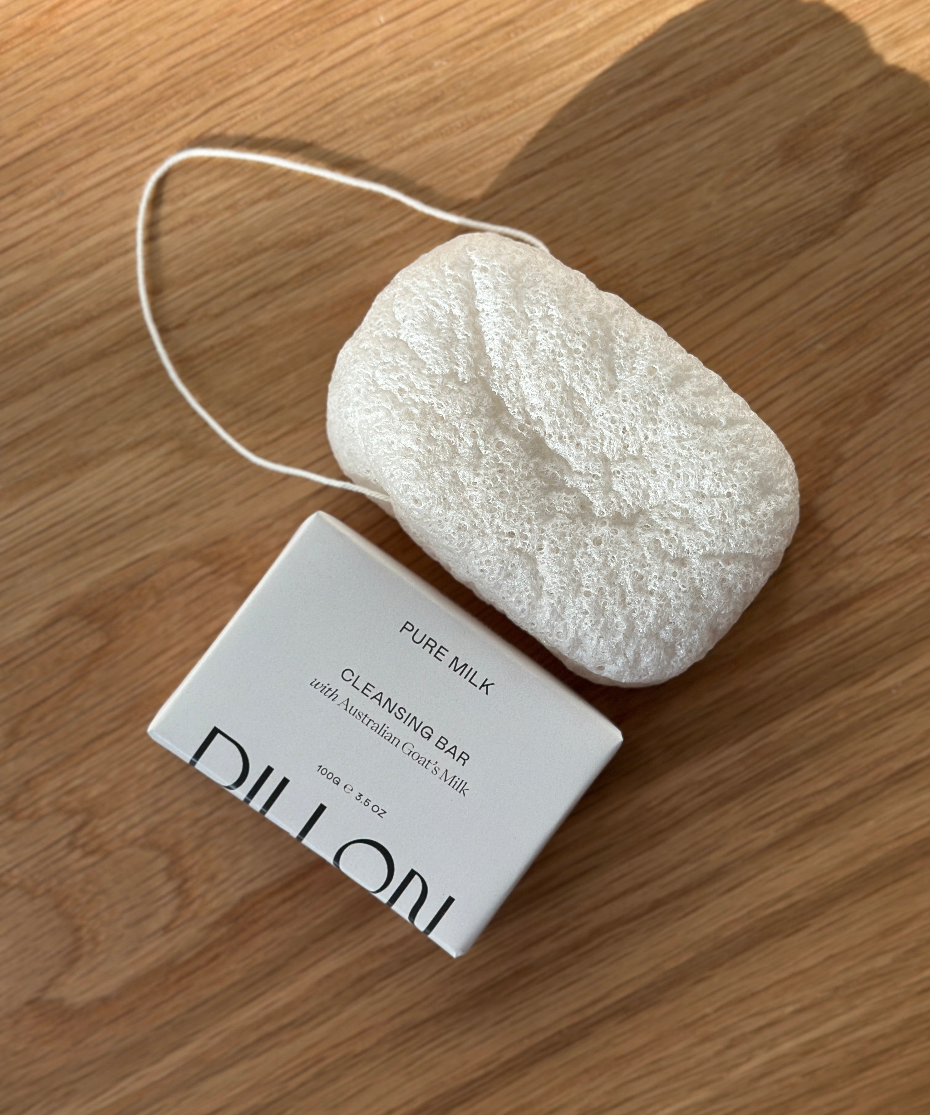 A white textured konjac body cleansing sponge next to bar soap in white paper packaging, labeled 'DILLON PURE MILK CLEANSING BAR', placed on a light brown wooden surface.