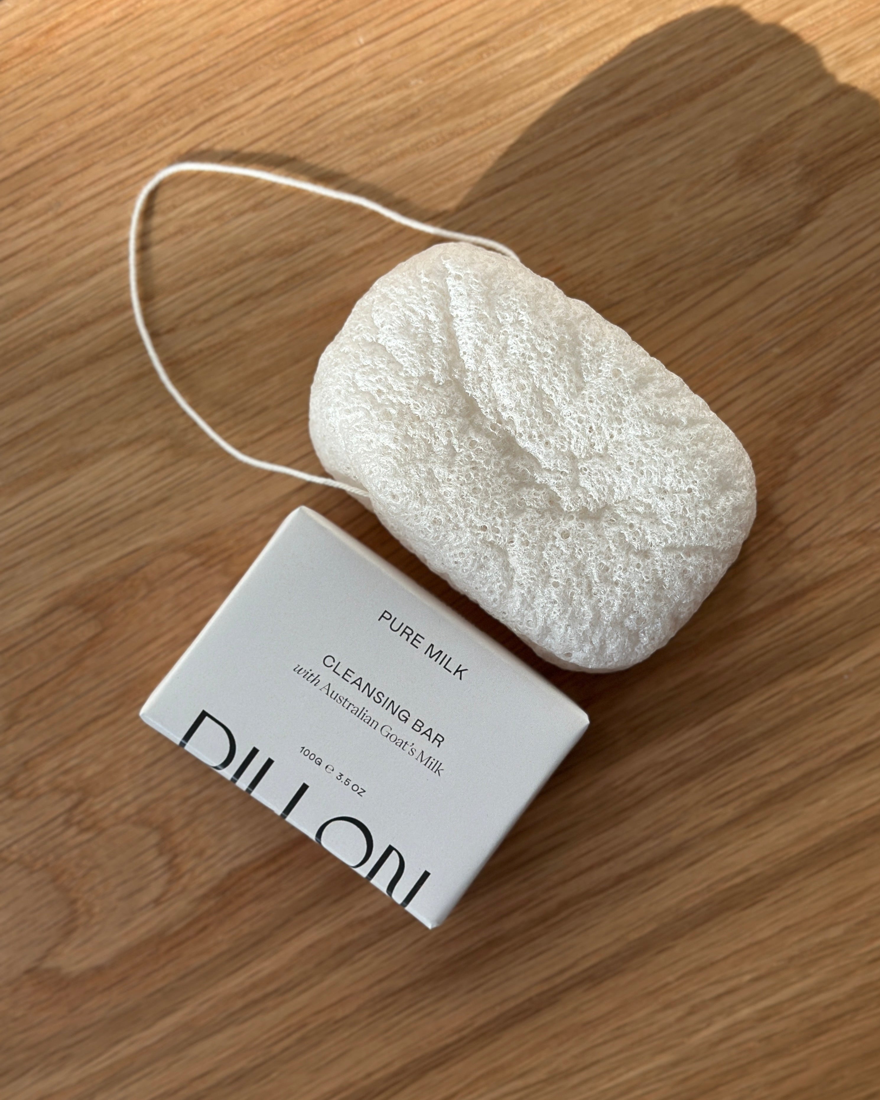 A white textured konjac body cleansing sponge next to bar soap in white paper packaging, labeled 'DILLON PURE MILK CLEANSING BAR', placed on a light brown wooden surface.