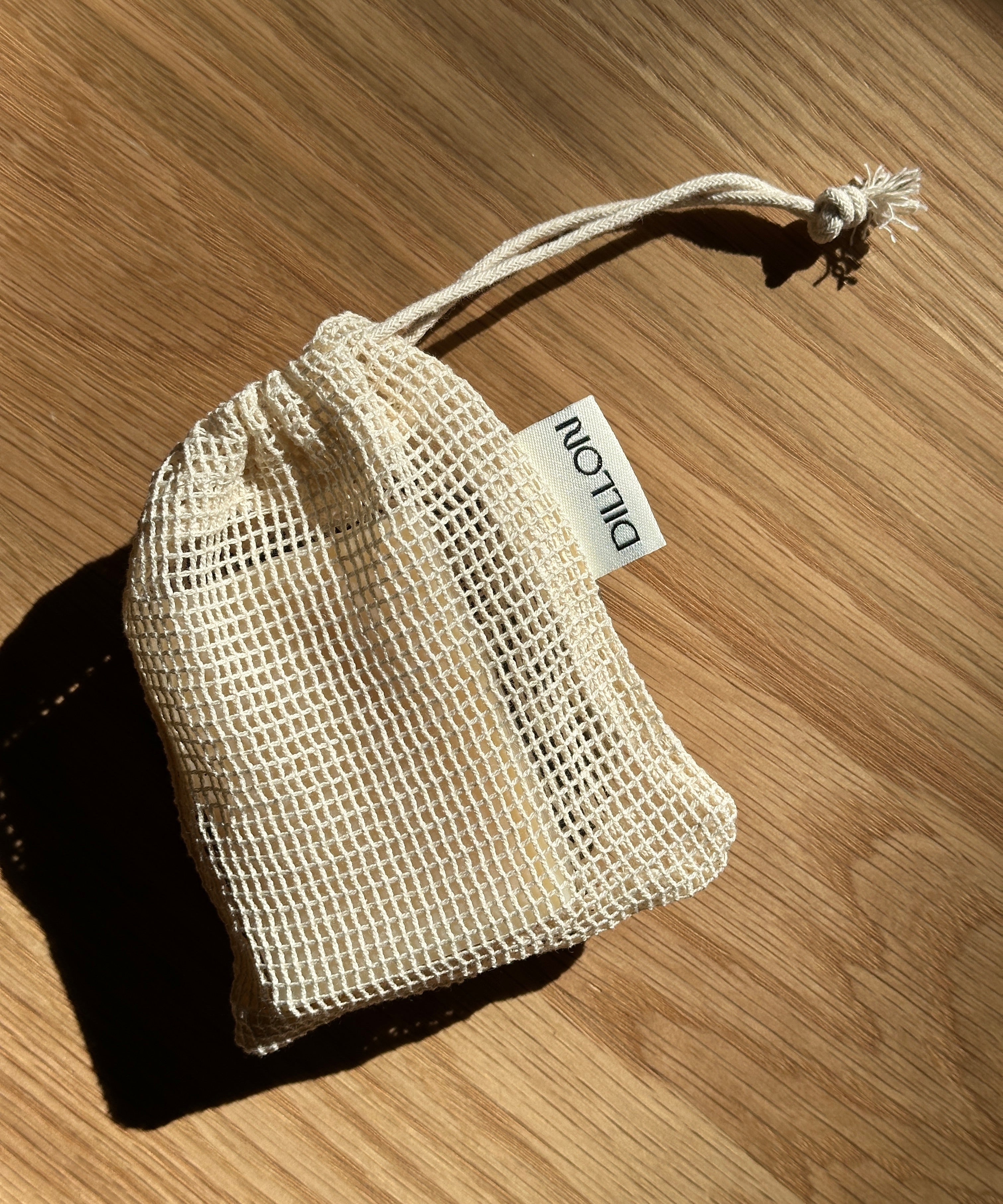A small cream-colored mesh drawstring pouch with a white tag reading 'DILLON' placed on a wooden surface.