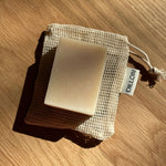 A rectangular bar of soap in a beige mesh drawstring pouch, photographed on a wooden surface with strong sunlight and shadows. A small tag on the pouch reads DILLON.