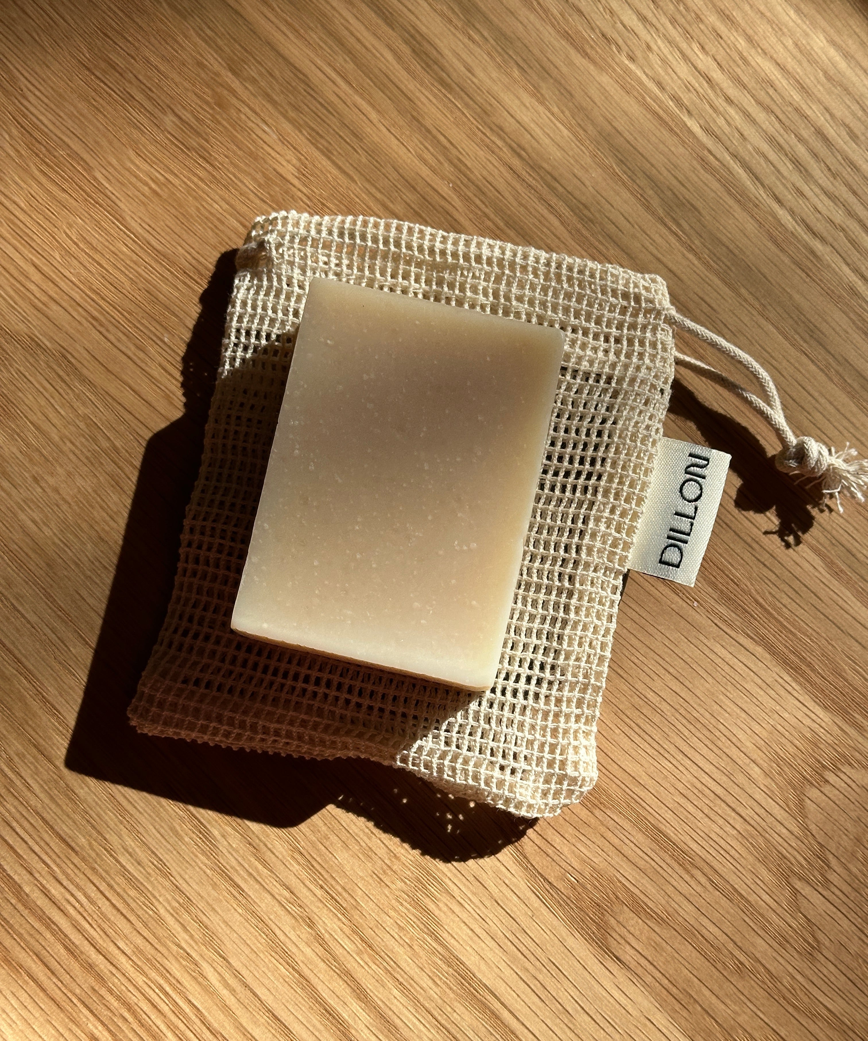 A rectangular bar of soap in a beige mesh drawstring pouch, photographed on a wooden surface with strong sunlight and shadows. A small tag on the pouch reads DILLON.