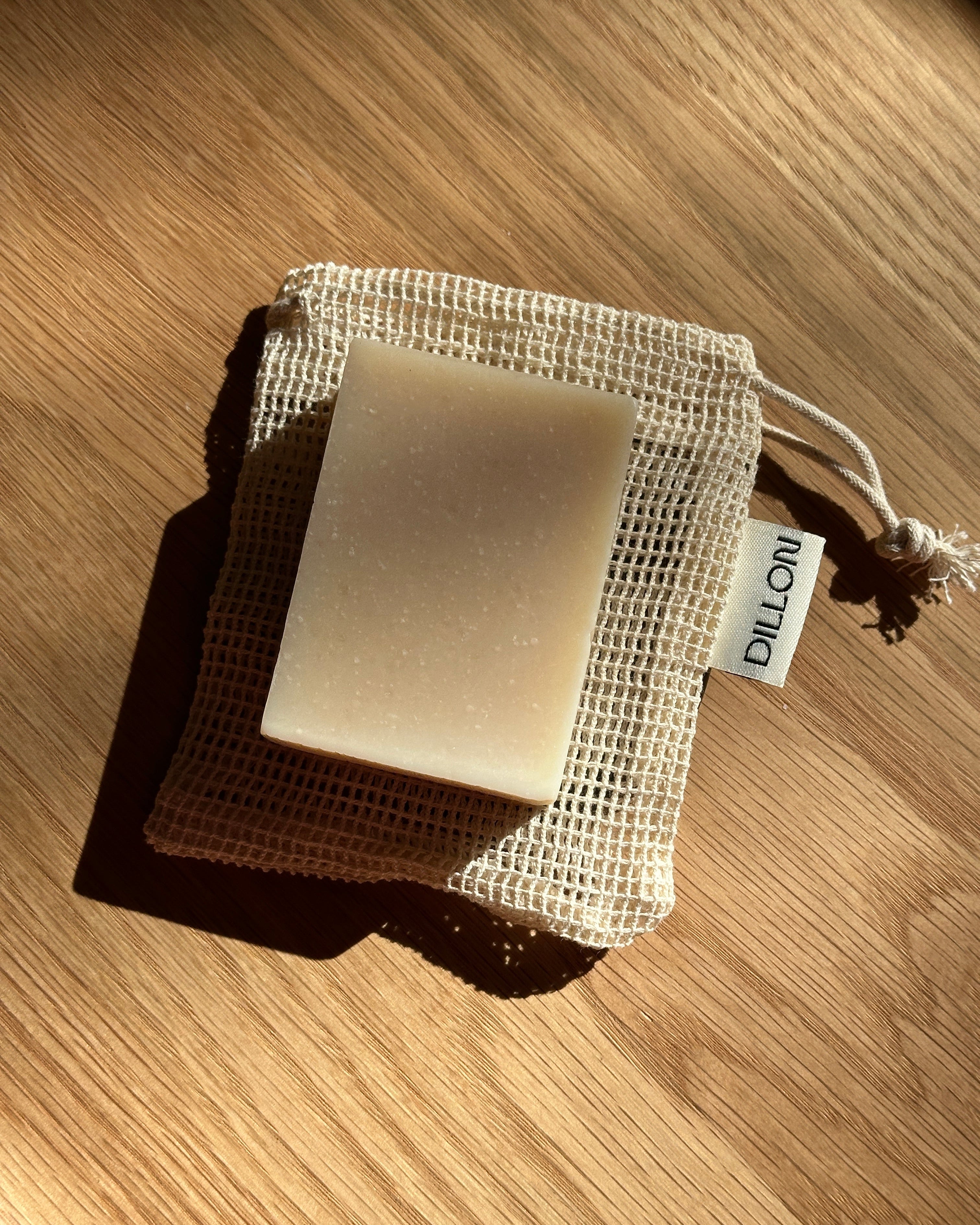 A rectangular bar of soap in a beige mesh drawstring pouch, photographed on a wooden surface with strong sunlight and shadows. A small tag on the pouch reads DILLON.
