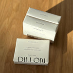Three white rectangular boxes of Dillon Pure Milk Cleansing Bar soap arranged on a wooden tabletop, with one box in front and two stacked behind it. The front box reads 'DILLON' and 'PURE MILK CLEANSING BAR,' while the boxes behind read 'SOOTHING' and 'PURE MILK.
