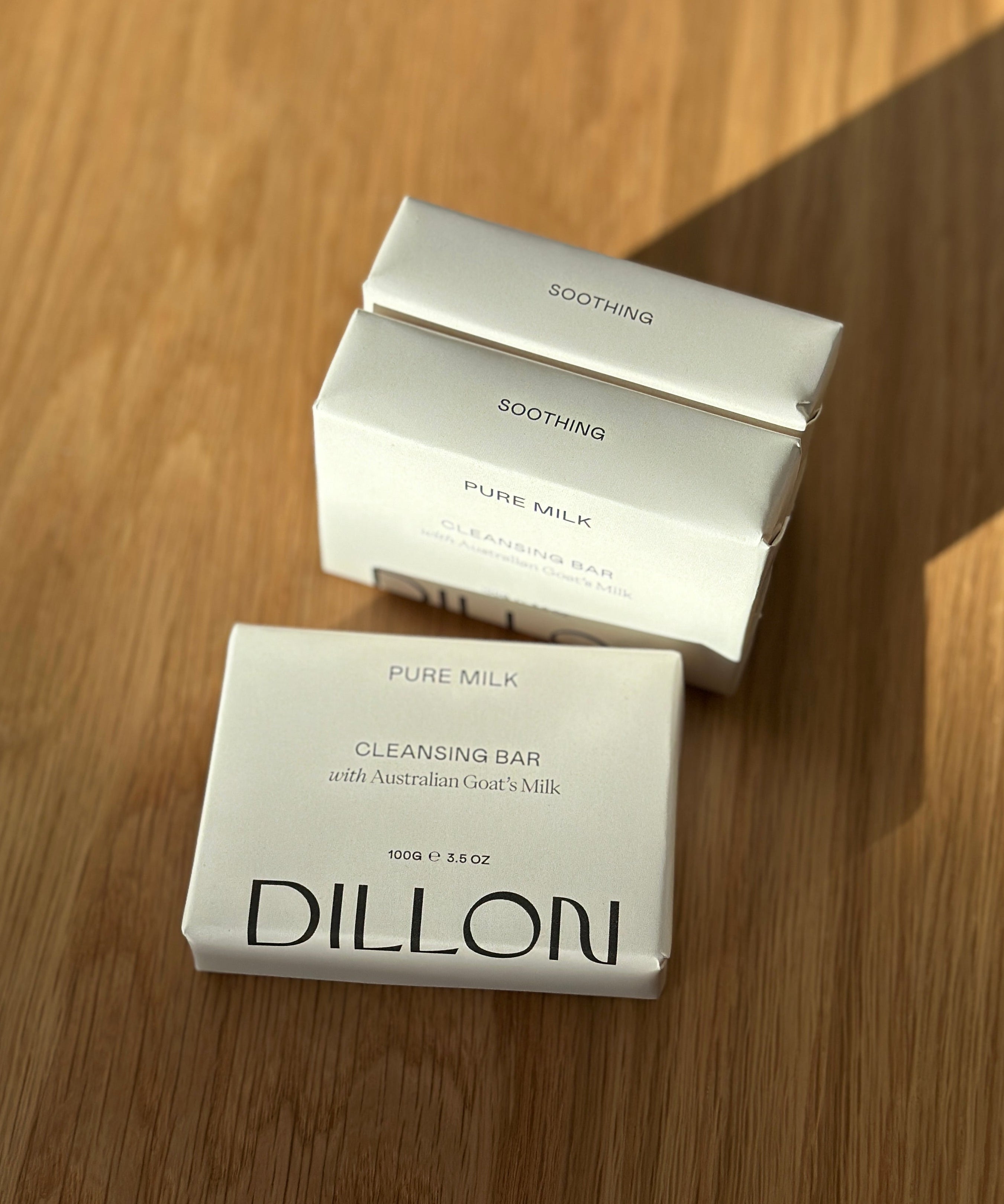 Three white rectangular boxes of Dillon Pure Milk Cleansing Bar soap arranged on a wooden tabletop, with one box in front and two stacked behind it. The front box reads 'DILLON' and 'PURE MILK CLEANSING BAR,' while the boxes behind read 'SOOTHING' and 'PURE MILK.