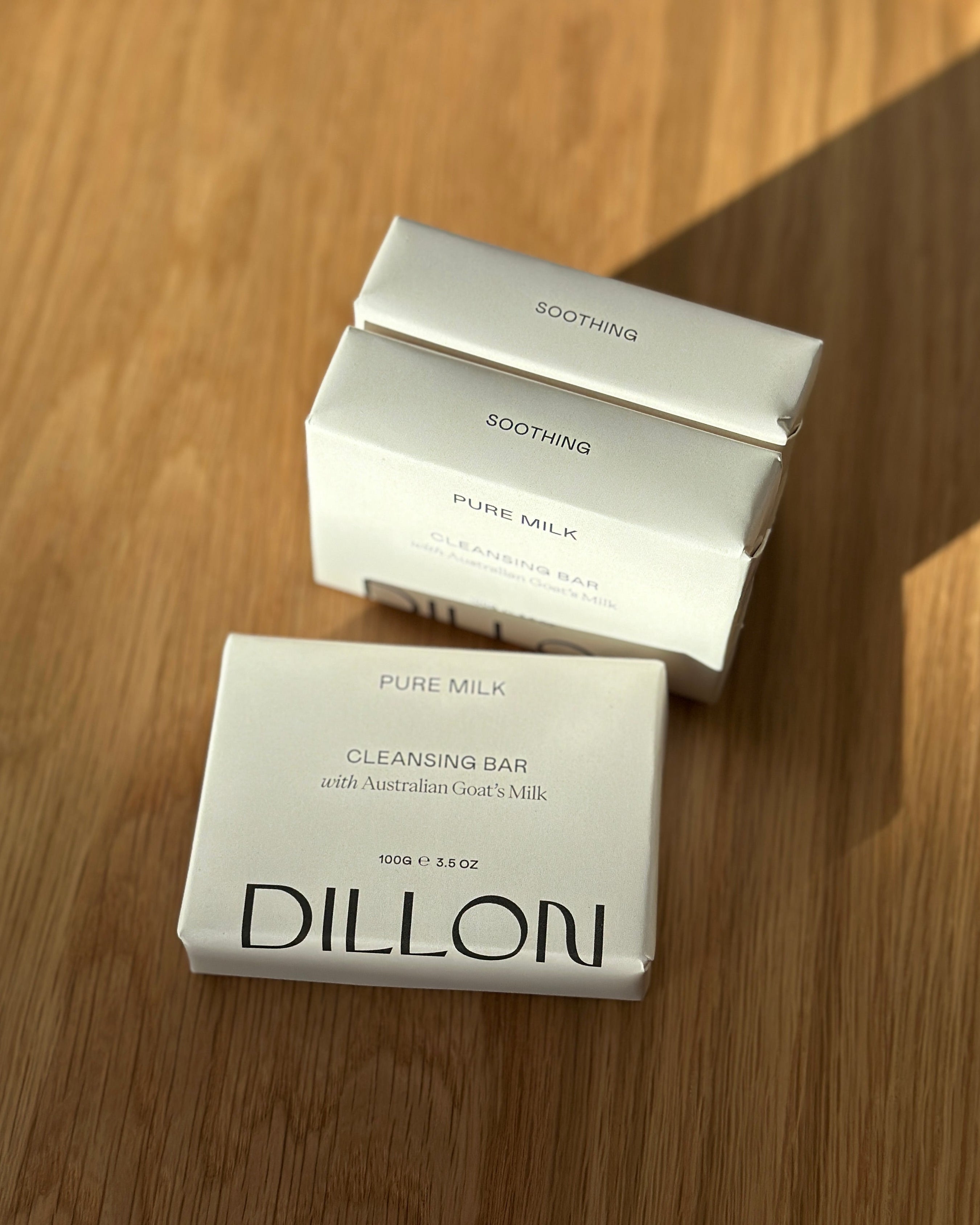 Three white rectangular boxes of Dillon Pure Milk Cleansing Bar soap arranged on a wooden tabletop, with one box in front and two stacked behind it. The front box reads 'DILLON' and 'PURE MILK CLEANSING BAR,' while the boxes behind read 'SOOTHING' and 'PURE MILK.