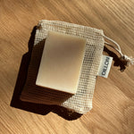 A rectangular bar of soap in a beige mesh drawstring pouch, photographed on a wooden surface with strong sunlight and shadows. A small tag on the pouch reads DILLON.