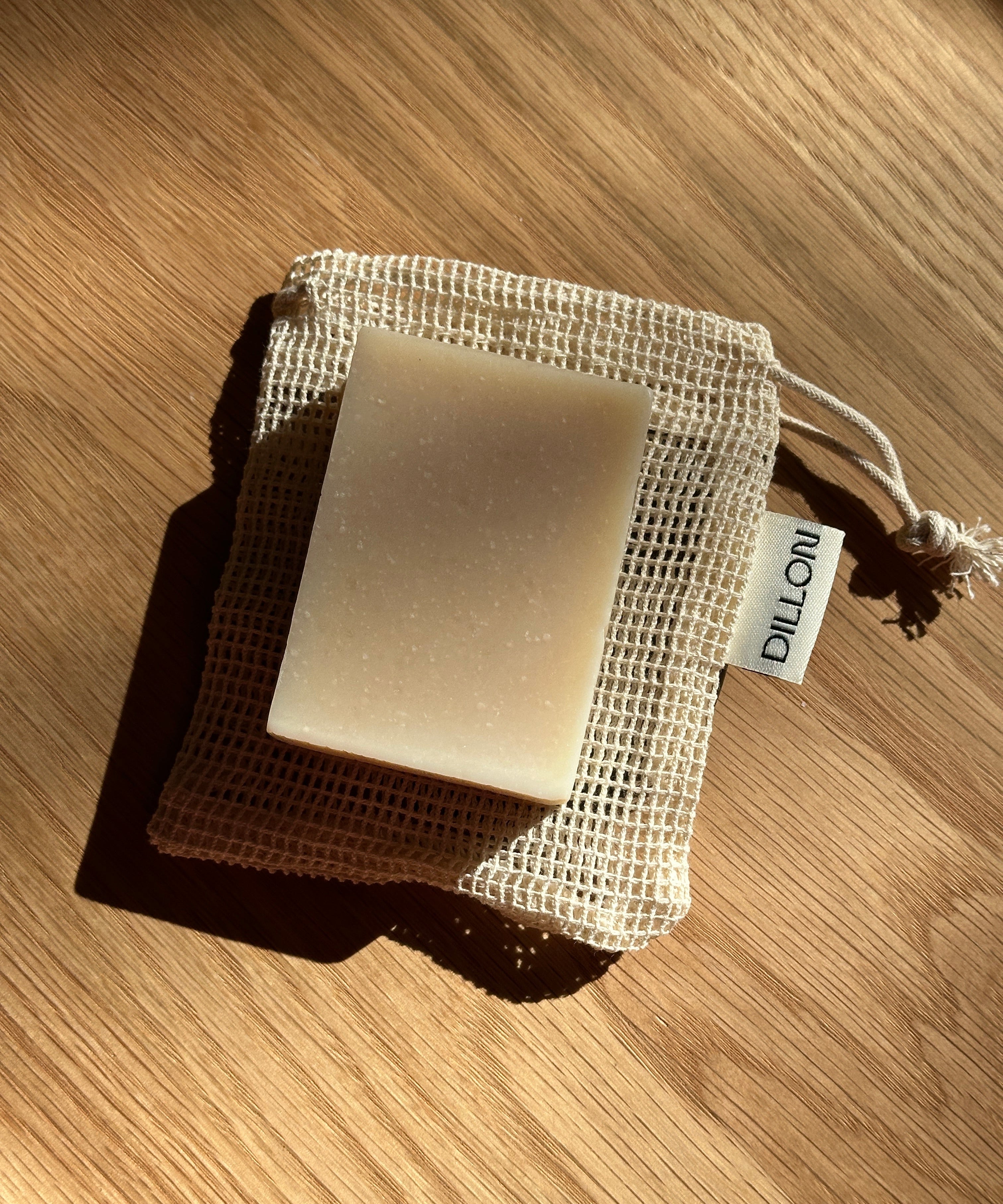 A rectangular bar of soap in a beige mesh drawstring pouch, photographed on a wooden surface with strong sunlight and shadows. A small tag on the pouch reads DILLON.