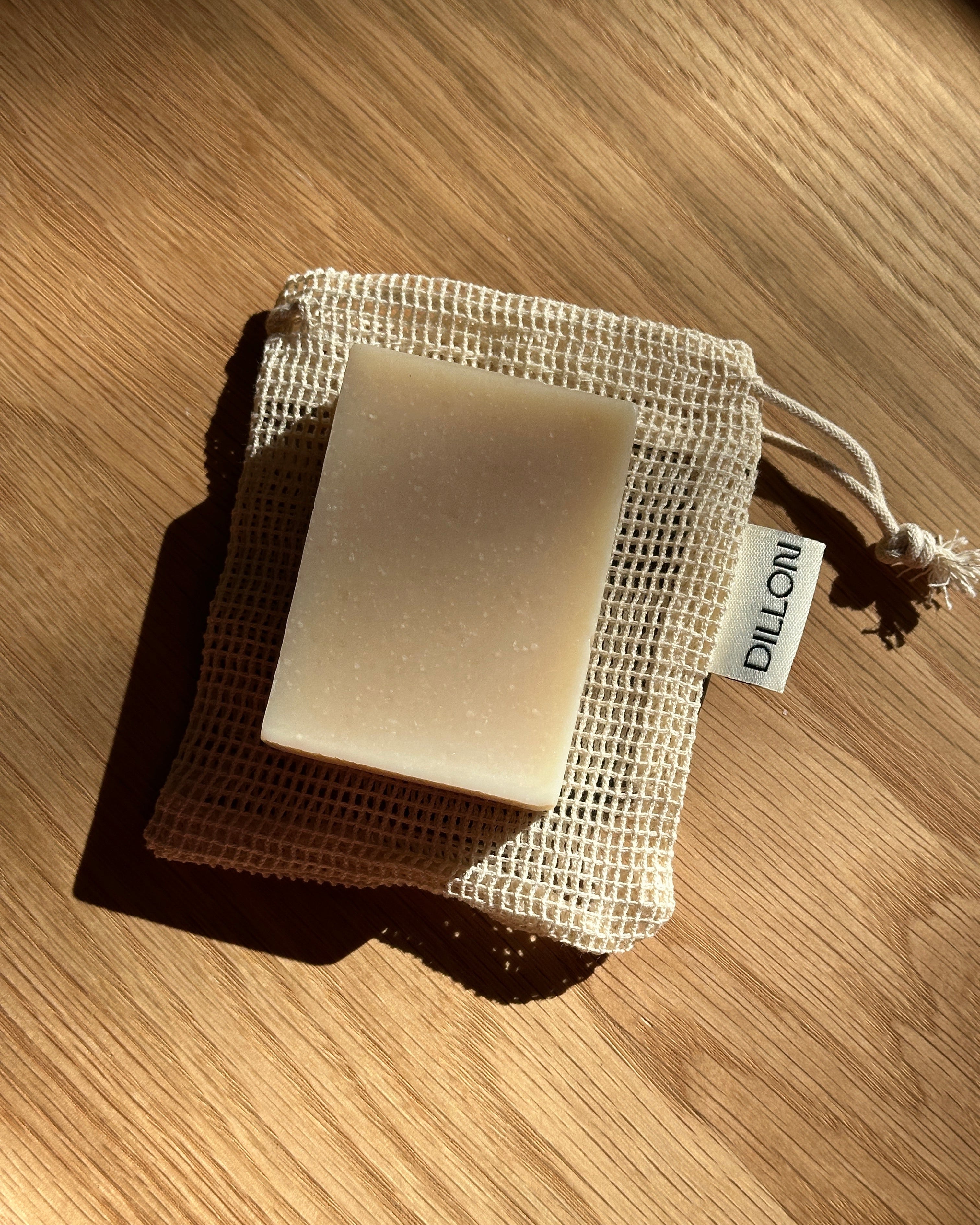 A rectangular bar of soap in a beige mesh drawstring pouch, photographed on a wooden surface with strong sunlight and shadows. A small tag on the pouch reads DILLON.