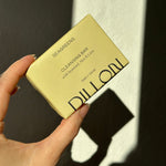 A hand holding a pale yellow DILLON Seagreens Cleansing Bar, set against a dark wall with strong sunlight and shadows.