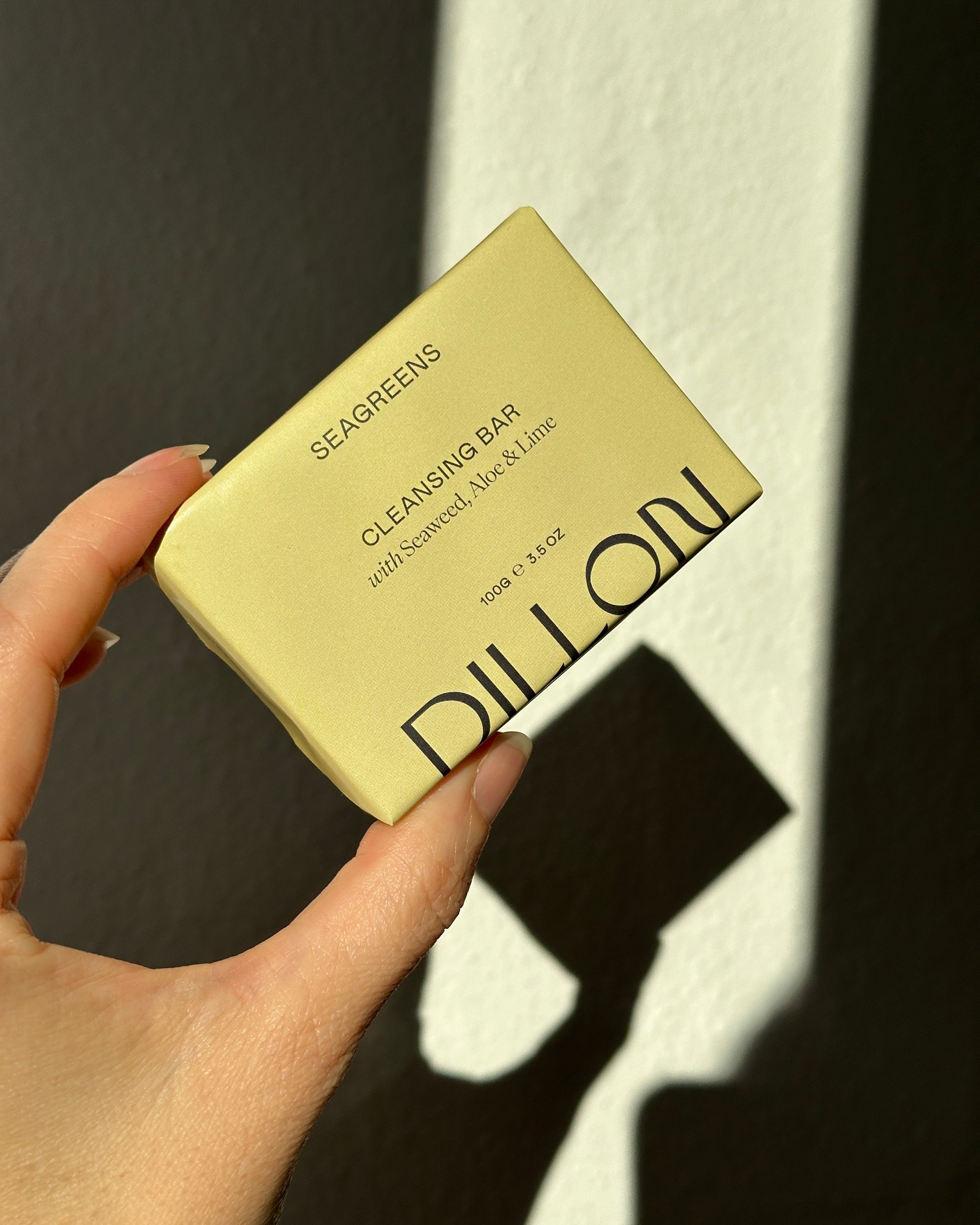 A hand holding a pale yellow DILLON Seagreens Cleansing Bar, set against a dark wall with strong sunlight and shadows.