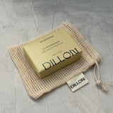 A bar of Dillon soap in a beige mesh drawstring pouch on a textured gray surface.