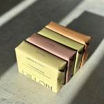 Four rectangular bar soaps stacked diagonally on a stone-like surface, each in a different metallic color: pale yellow, dusty rose, olive green, and copper. The front box reads 'DILLON' and 'LEMON & HONEY CLEANSING BAR'.
