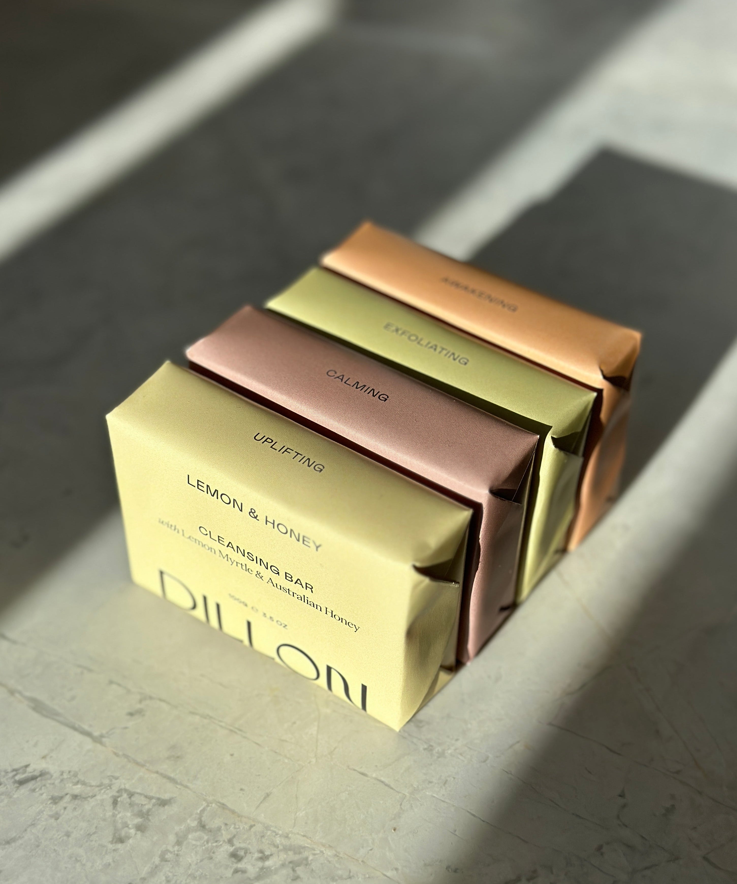 Four rectangular bar soaps stacked diagonally on a stone-like surface, each in a different metallic color: pale yellow, dusty rose, olive green, and copper. The front box reads 'DILLON' and 'LEMON & HONEY CLEANSING BAR'.