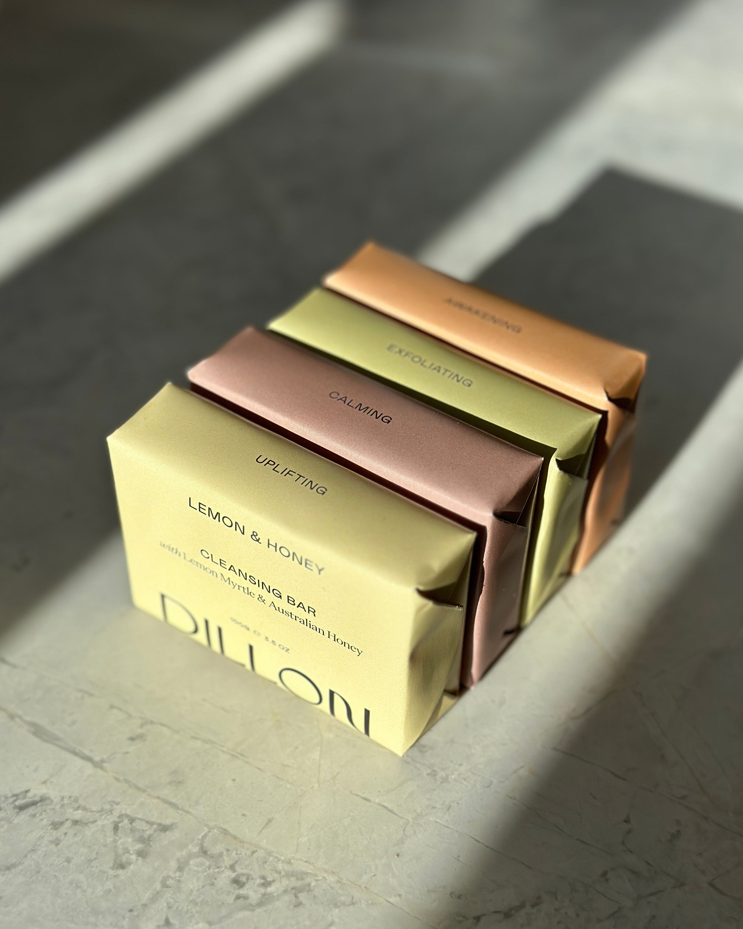Four rectangular bar soaps stacked diagonally on a stone-like surface, each in a different metallic color: pale yellow, dusty rose, olive green, and copper. The front box reads 'DILLON' and 'LEMON & HONEY CLEANSING BAR'.